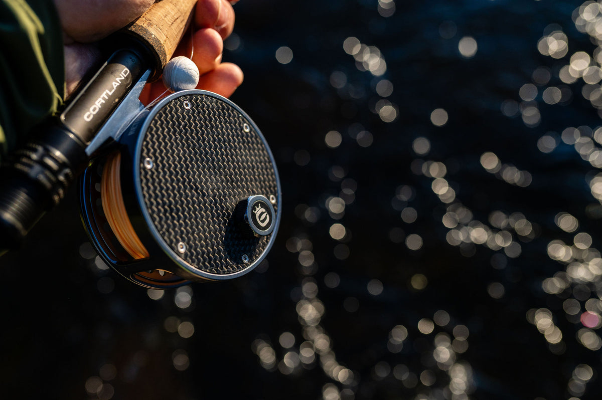 Close-up of a fly fishing reel with a carbon fiber patterned spool, held by a person whose hand is visible, with a blurred background of sparkling water.